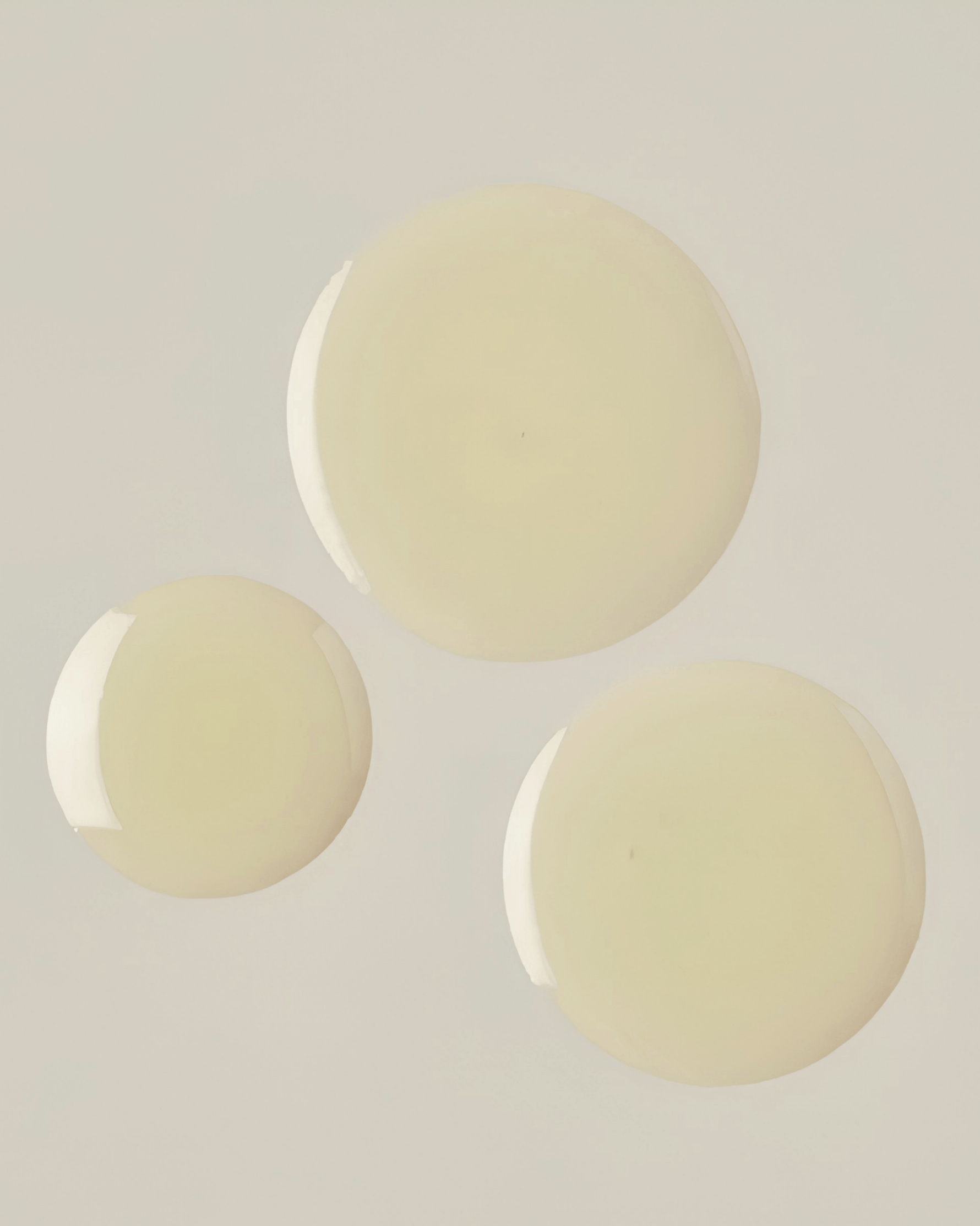 Three pale yellow oil droplets of face serum on a neutral background